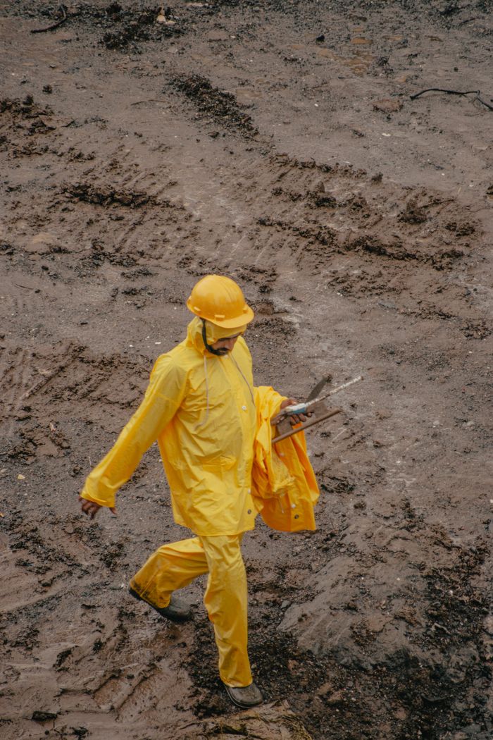 services-02 Worker in yellow raincoat on muddy construction site carrying tools, safety-focused.